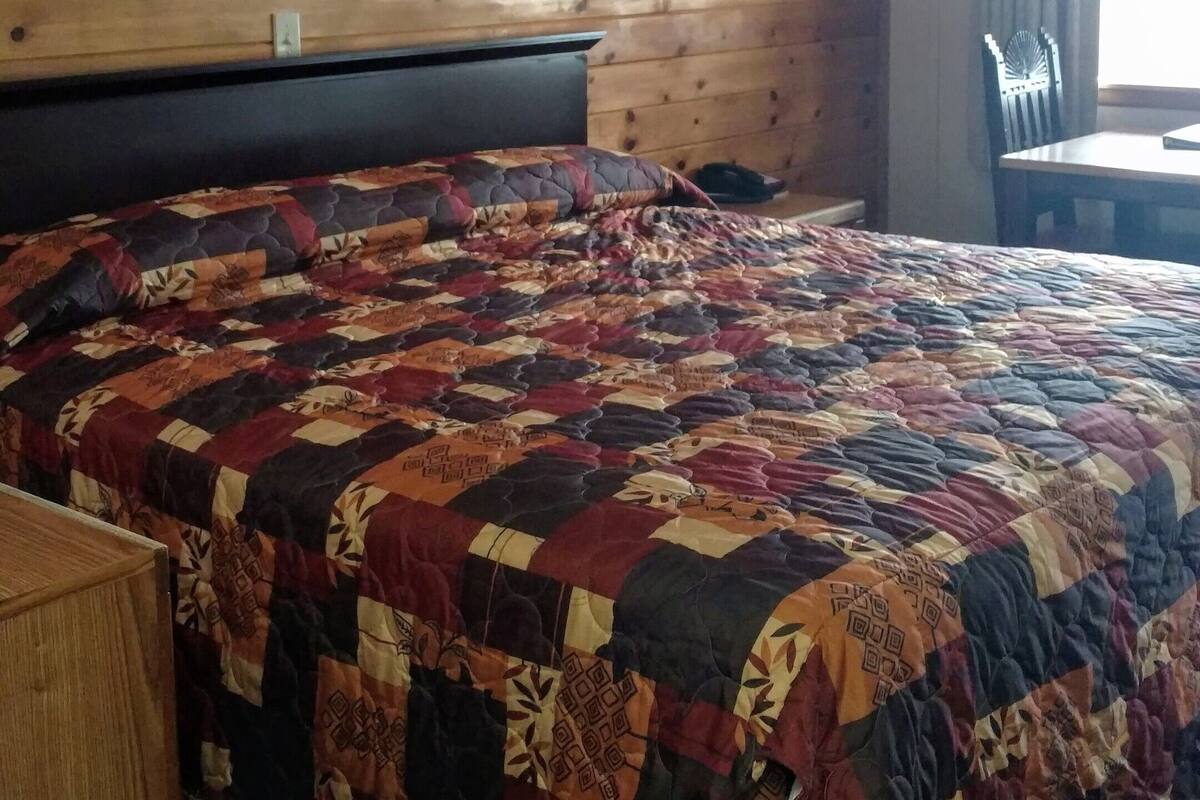 A hotel room with a wooden headboard, a large bed with a colorful quilt, a bedside table, a chair, and a window with curtains.
