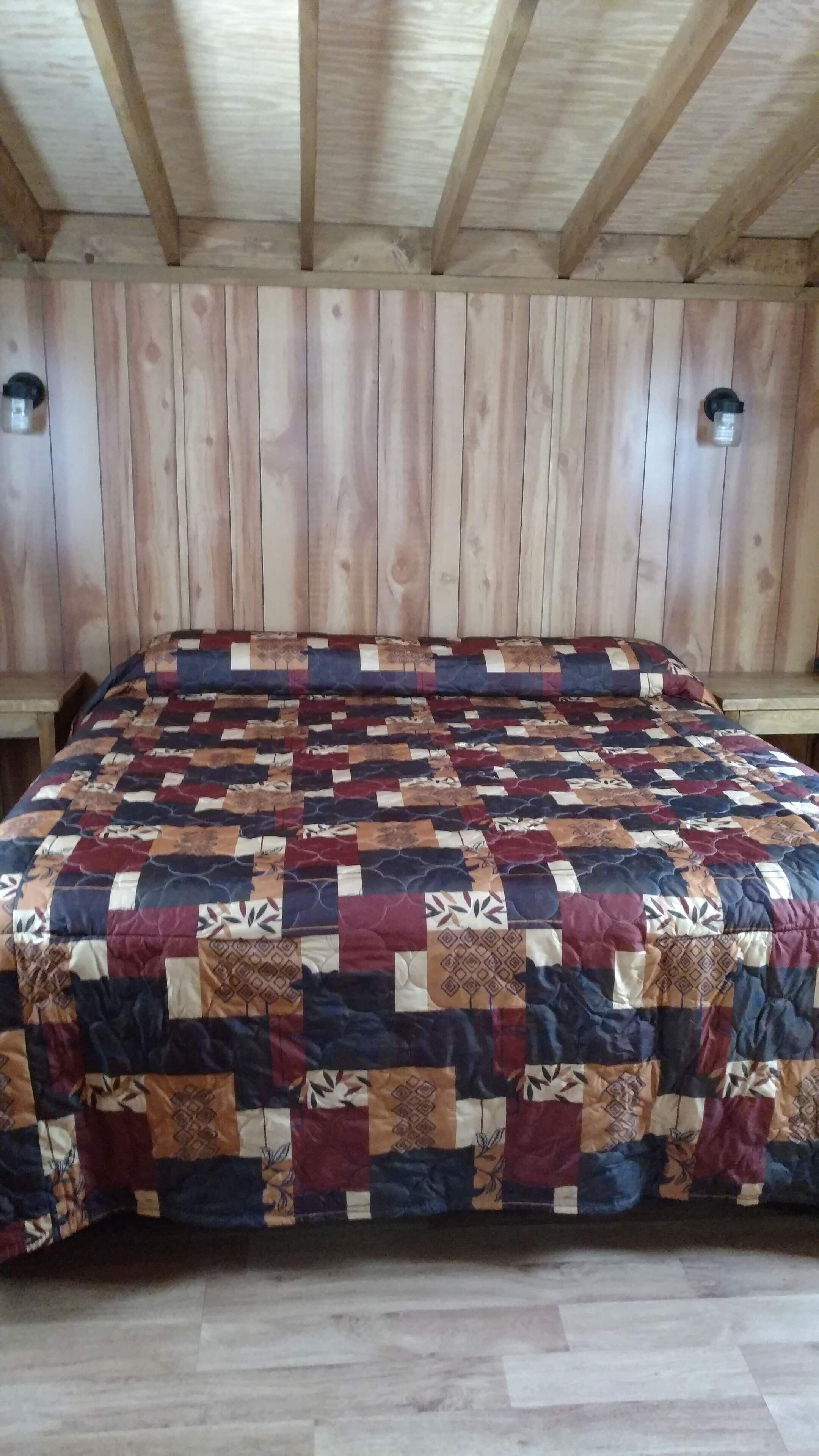 Une pièce avec un plafond et des murs en bois, un lit recouvert d’une couverture aux couleurs vives, et deux appliques murales.