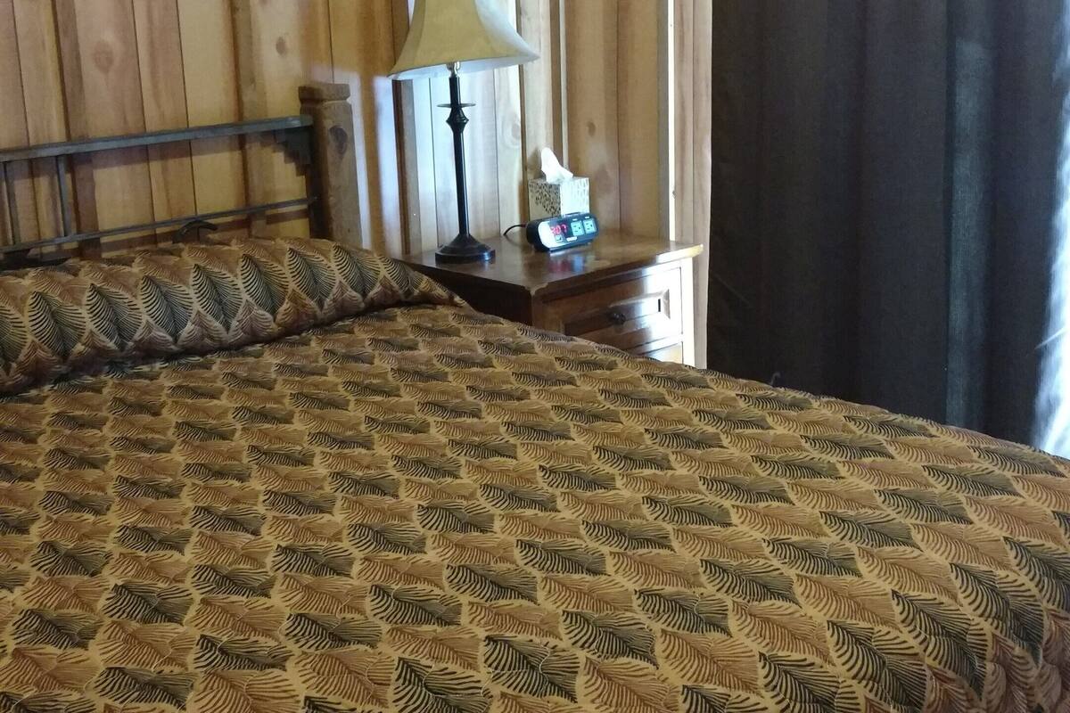 A bedroom with a patterned bedspread, a bedside lamp, a clock, and a curtain.