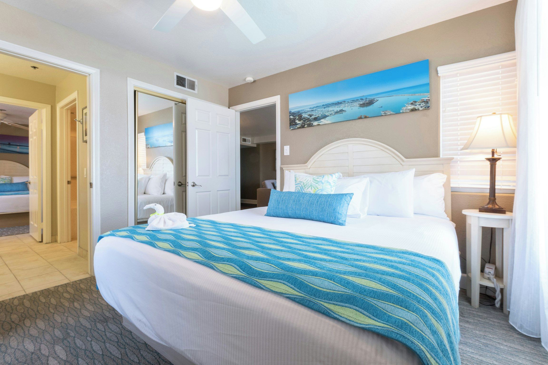 A hotel room with a large bed, a painting of a coastal scene, a bedside lamp, and a view of the adjacent room.