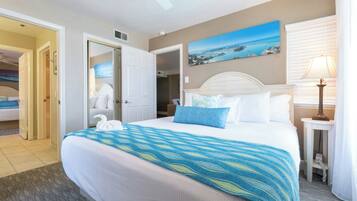 A hotel room with a large bed, a painting of a coastal scene, a bedside lamp, and a view of the adjacent room.