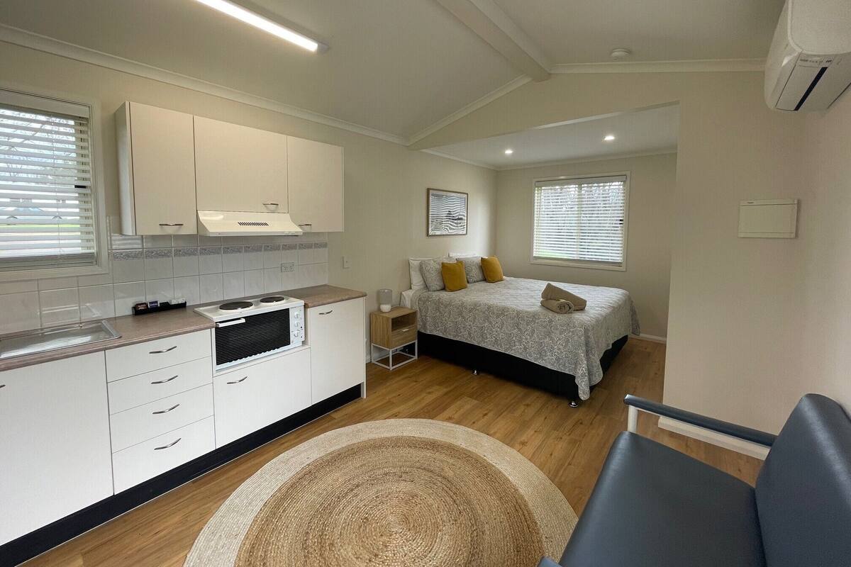 A compact hotel room with a kitchenette, a bed with two pillows, a bedside table, and a window with blinds.