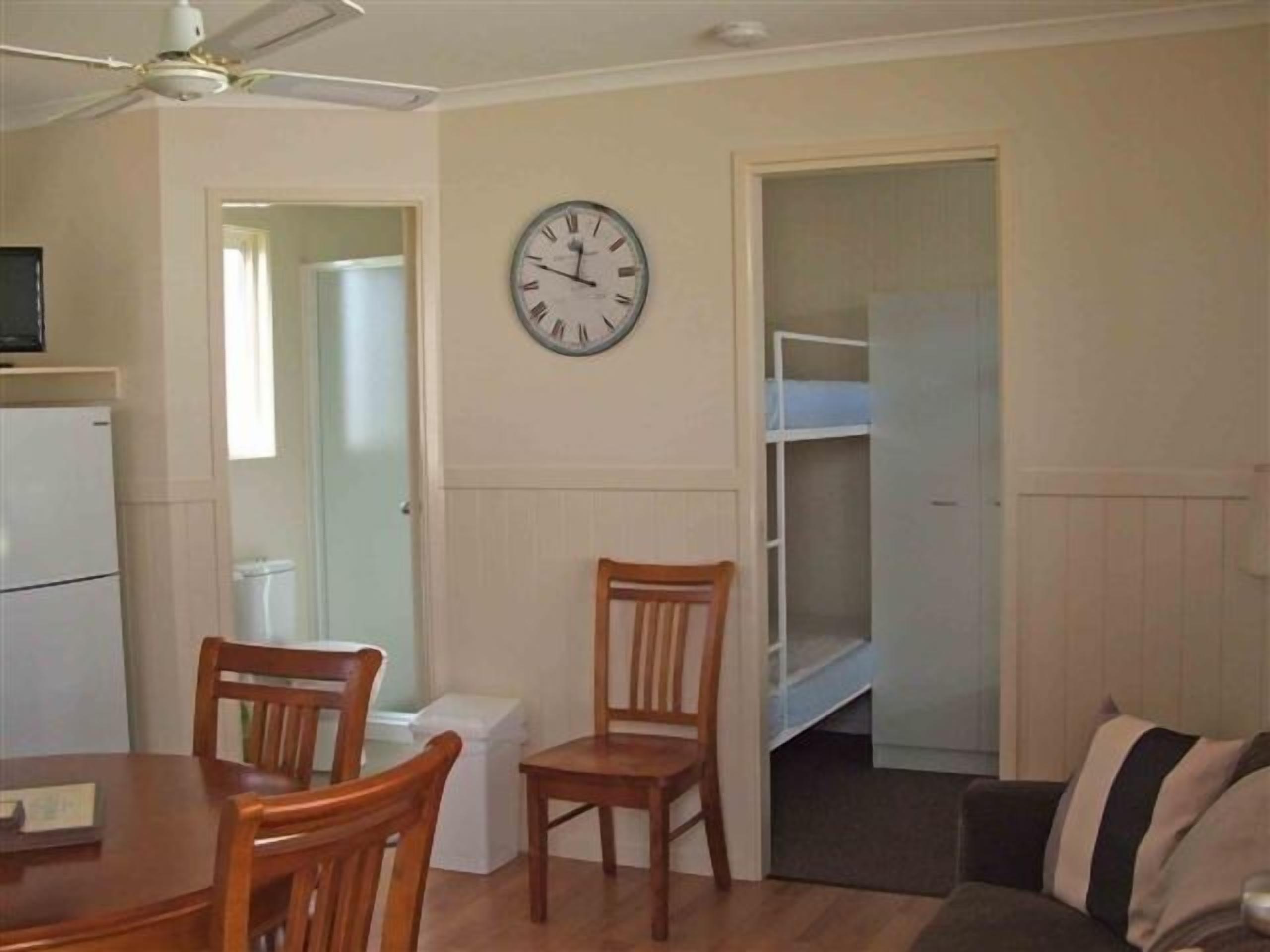 Una habitación con una mesa de comedor, sillas, un reloj, un ventilador de techo, una cama litera y un refrigerador.