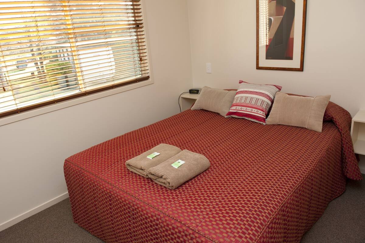 A neatly made bed with a red patterned bedspread, two folded towels, and two pillows on a white nightstand.