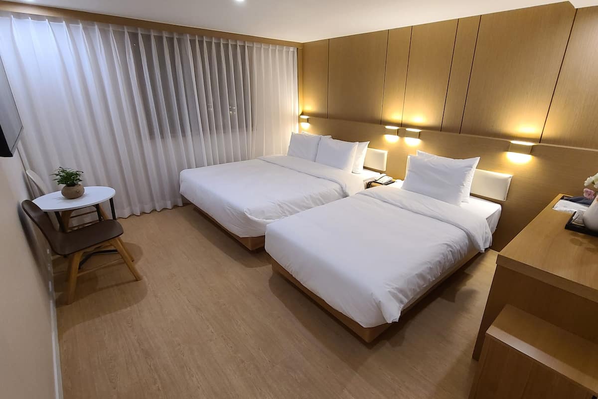 A hotel room with two beds, a small table with a plant, a desk with a vase of flowers, and a wooden headboard with wall-mounted lights.