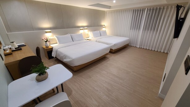 Ein Hotelzimmer mit zwei Betten, einem Schreibtisch, einem Stuhl, einem kleinen Tisch mit Pflanze und einem Fenster mit Vorhängen.