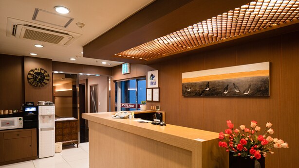 A hotel reception area with a wooden reception desk, a clock, a sign indicating 'Hotel Prime', a painting of birds, and a vase of flowers. - Hotel Prime Changwon (Changwon)