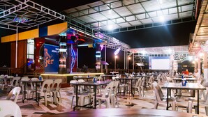 An indoor dining area with tables and chairs, a stage with a blue banner, and a large screen.