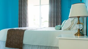 Una cama bien hecha con una colcha blanca y marrón, una lámpara de noche y una ventana con cortinas.