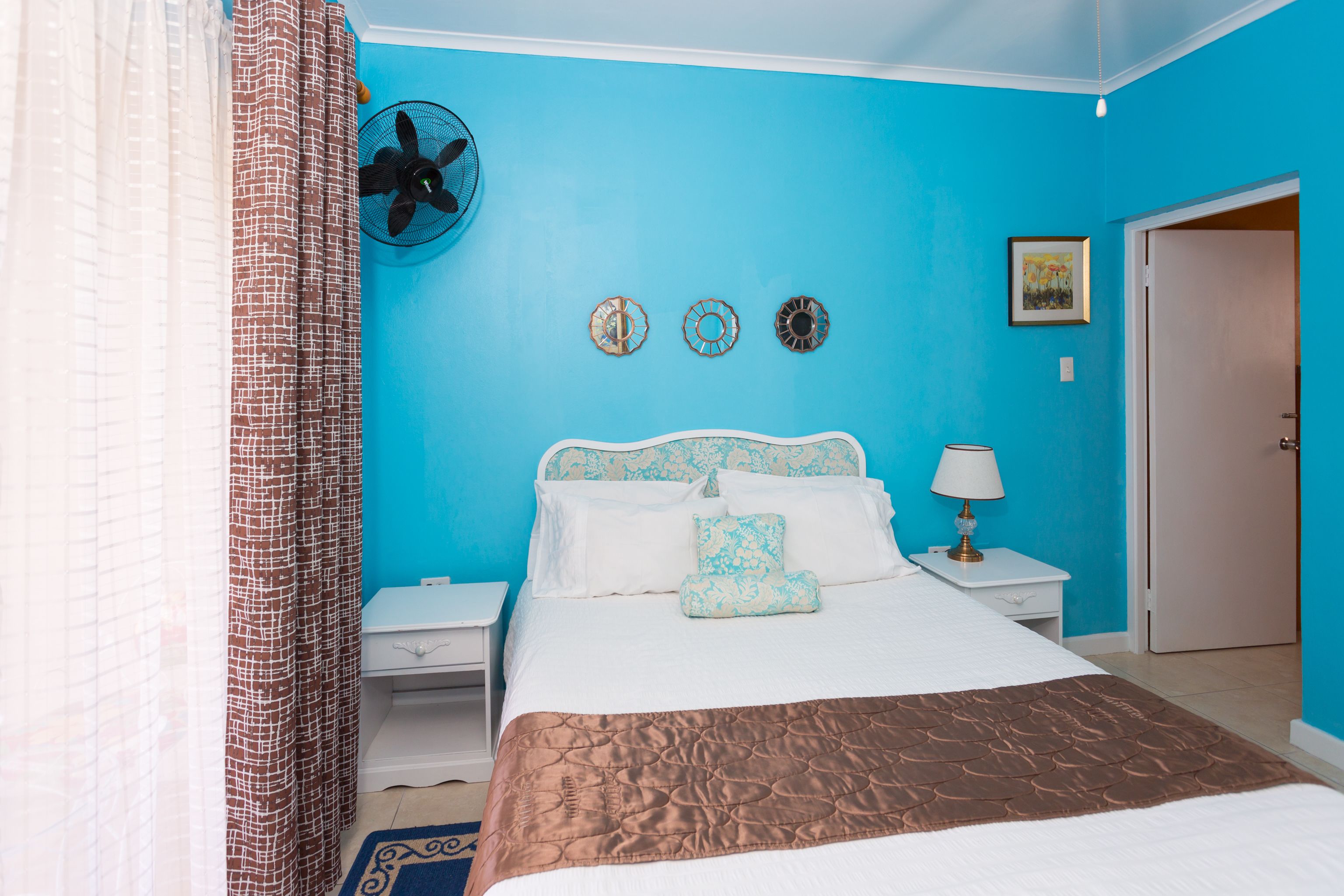 A bedroom with a blue wall, a bed with white and brown bedding, a nightstand with a lamp, and a fan on the wall.