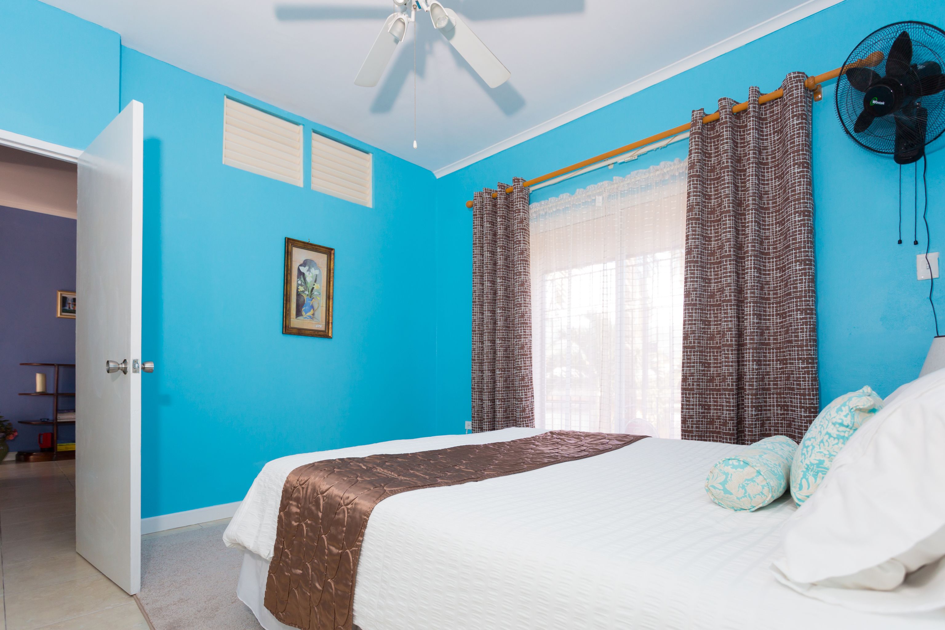 Une chambre à coucher avec un lit, un ventilateur de plafond, une fenêtre avec des rideaux et une porte.