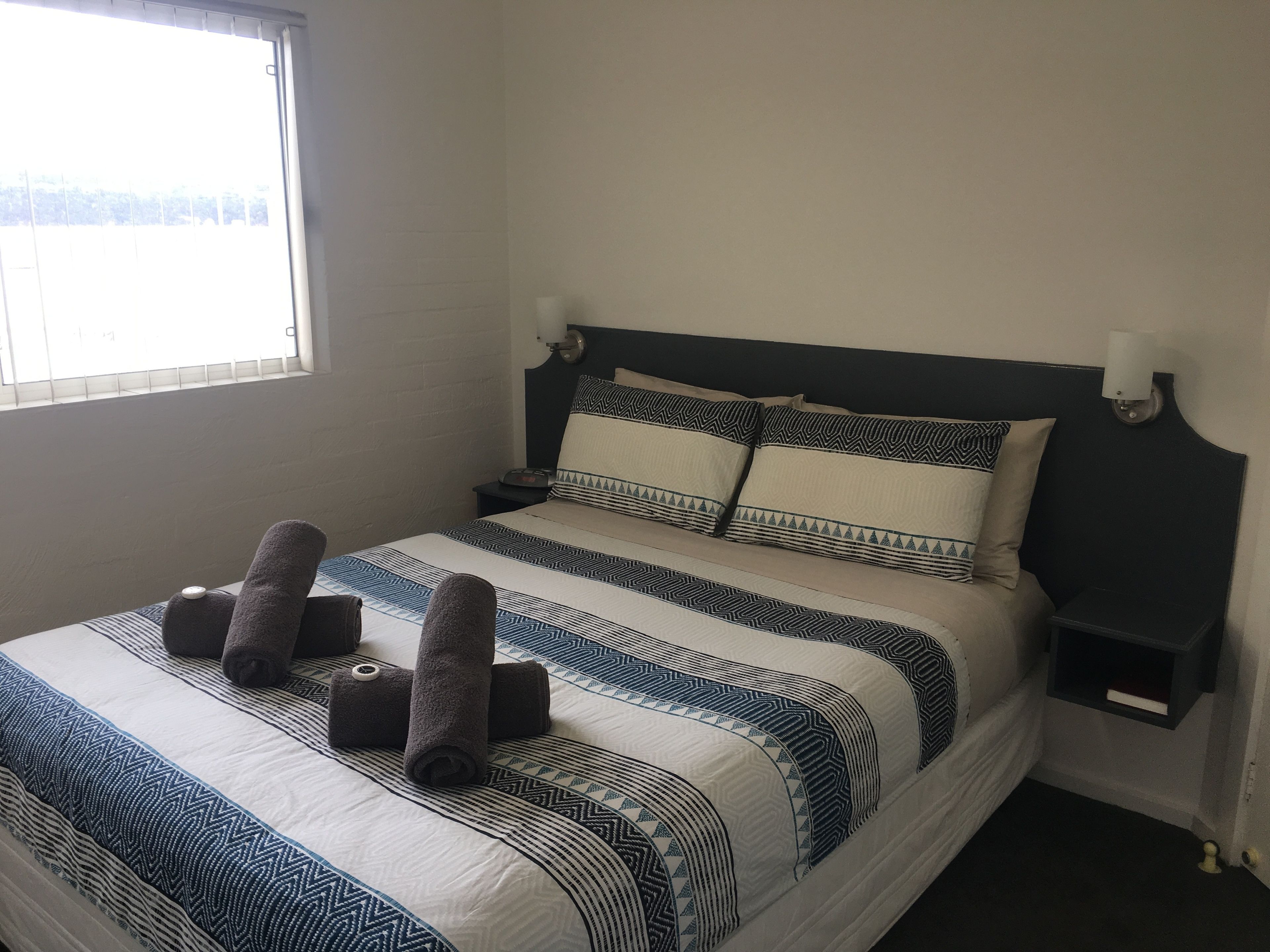 A neatly made bed with patterned bedding and rolled towels, a bedside table with a lamp, and a window with blinds.