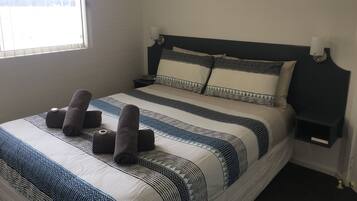 A neatly made bed with patterned bedding and rolled towels, a bedside table with a lamp, and a window with blinds.