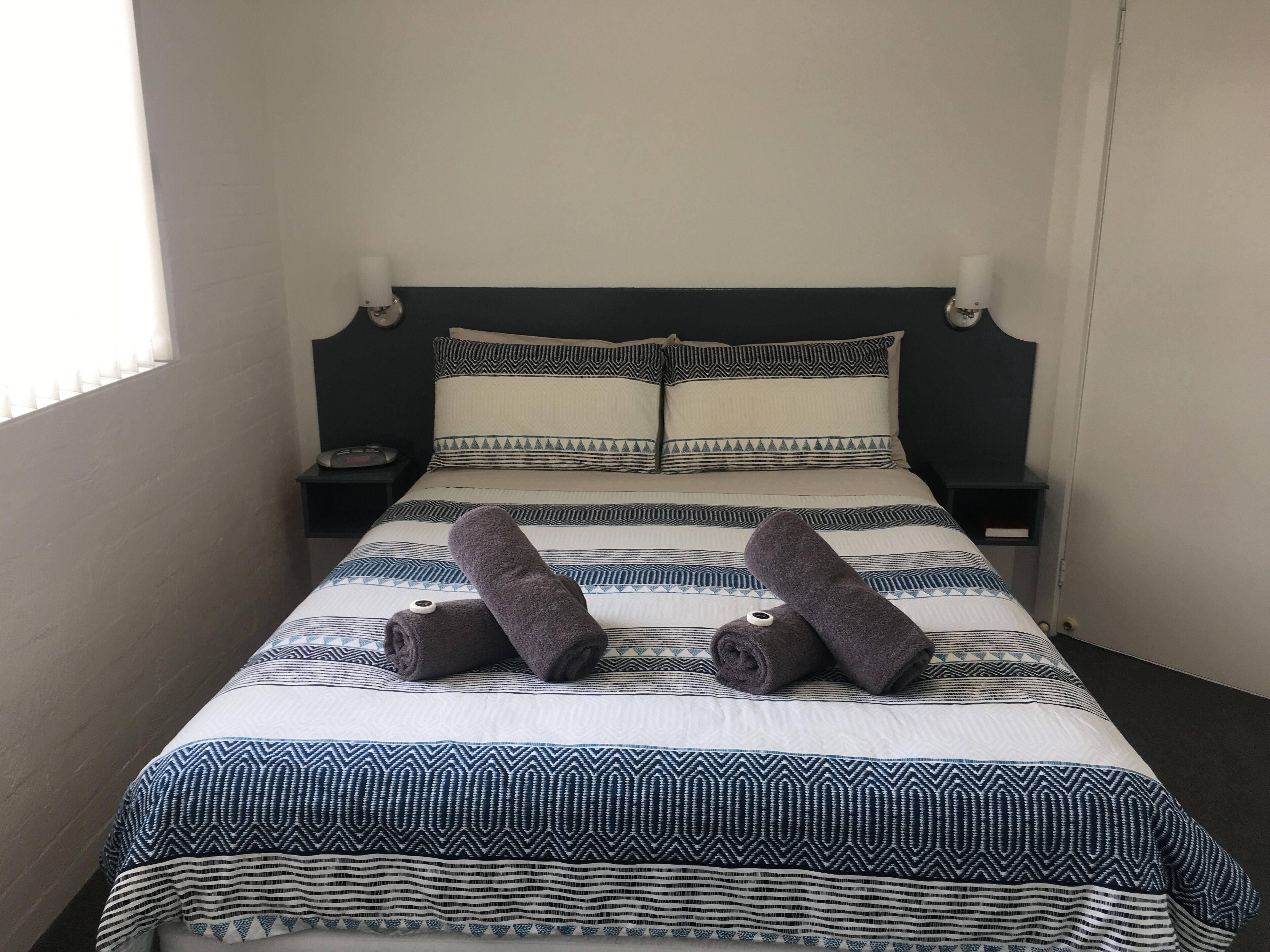 A neatly made bed with patterned bedding, two bedside tables, and a window with blinds.