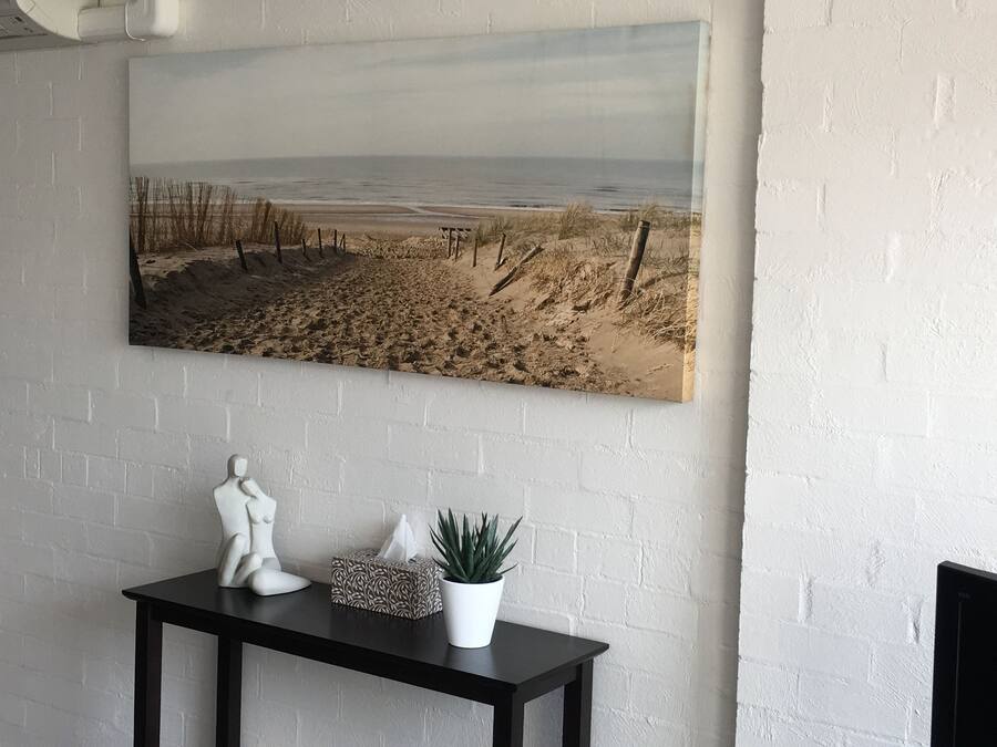 Pintura de una escena de playa sobre un muro de ladrillo blanco, una mesa pequeña con una planta, un adorno y una caja de pañuelos.