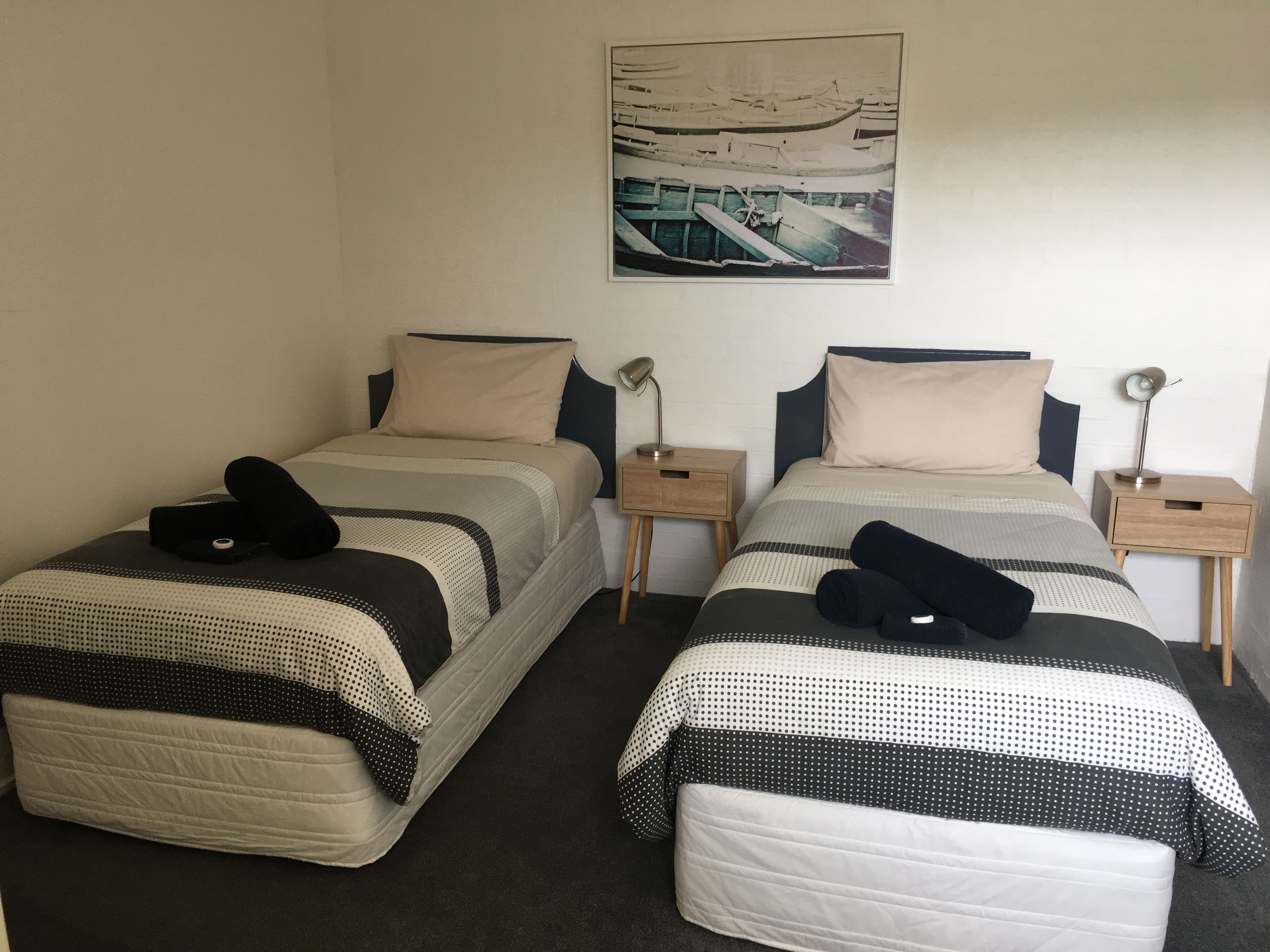 A room with two single beds, each with a pillow and a blanket, a nightstand with a lamp, and a framed picture on the wall.