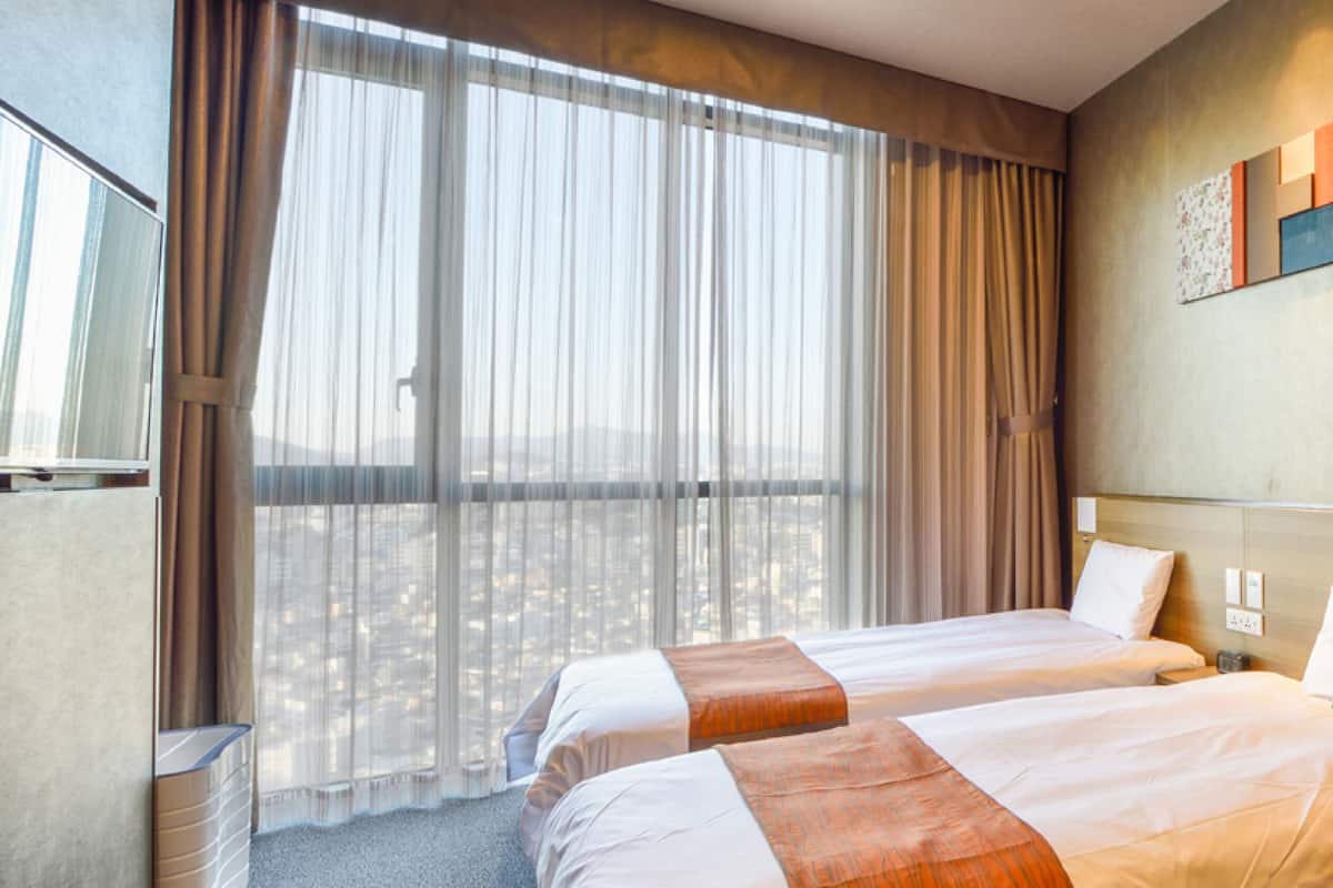 A hotel room with two beds, a large window with curtains, a wall-mounted artwork, and a seating area.