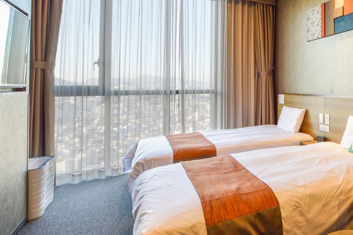 A hotel room with two beds, a sofa, a window with sheer curtains, and a city view.