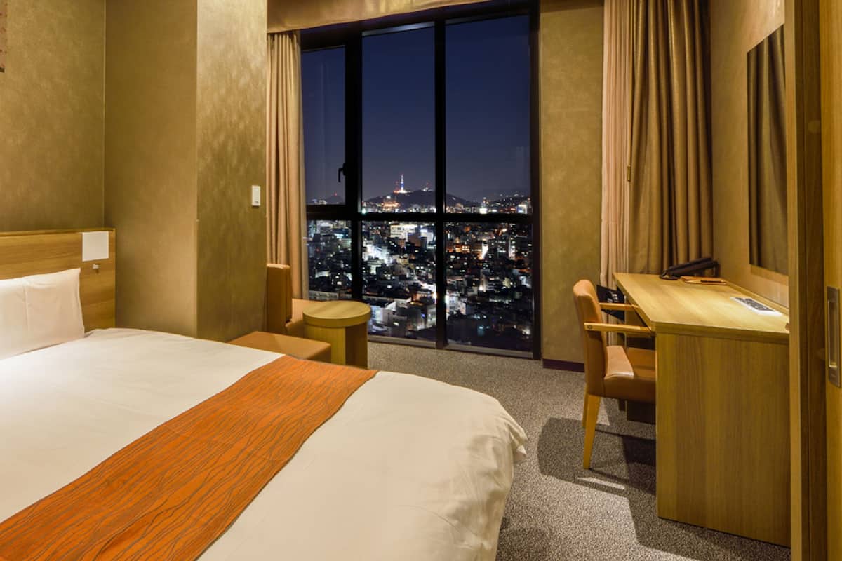 A hotel room with a large bed, a desk, and a view of a cityscape at night.
