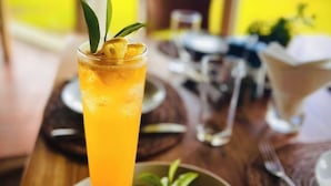 Una mesa dispuesta con un vaso alto de bebida de naranja decorado con una hoja y una rodaja de limón, un plato pequeño con una rodaja de limón, y un fondo desenfocado que sugiere una vista al aire libre.