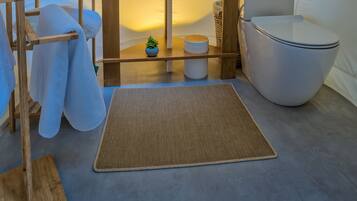 Habitación pequeña y luminosa con un espejo redondo, un lavamanos y un inodoro. Hay estantes de madera con toallas y una planta pequeña.