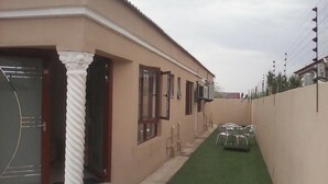 Un edificio con fachada color crema, una columna blanca y un césped artificial verde. Hay sillas y una pequeña mesa al aire libre.