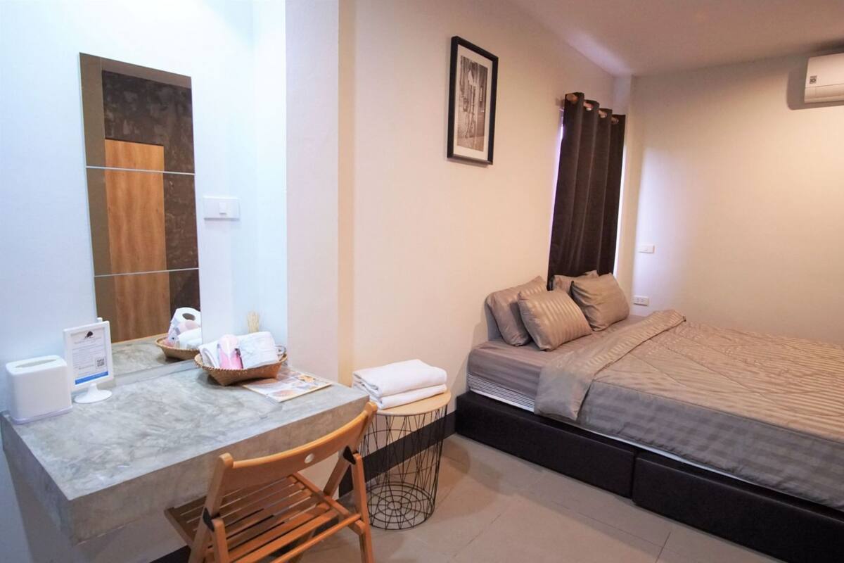 A compact hotel room with a bed, a small desk with a chair, a trash bin, and a wall-mounted air conditioner.