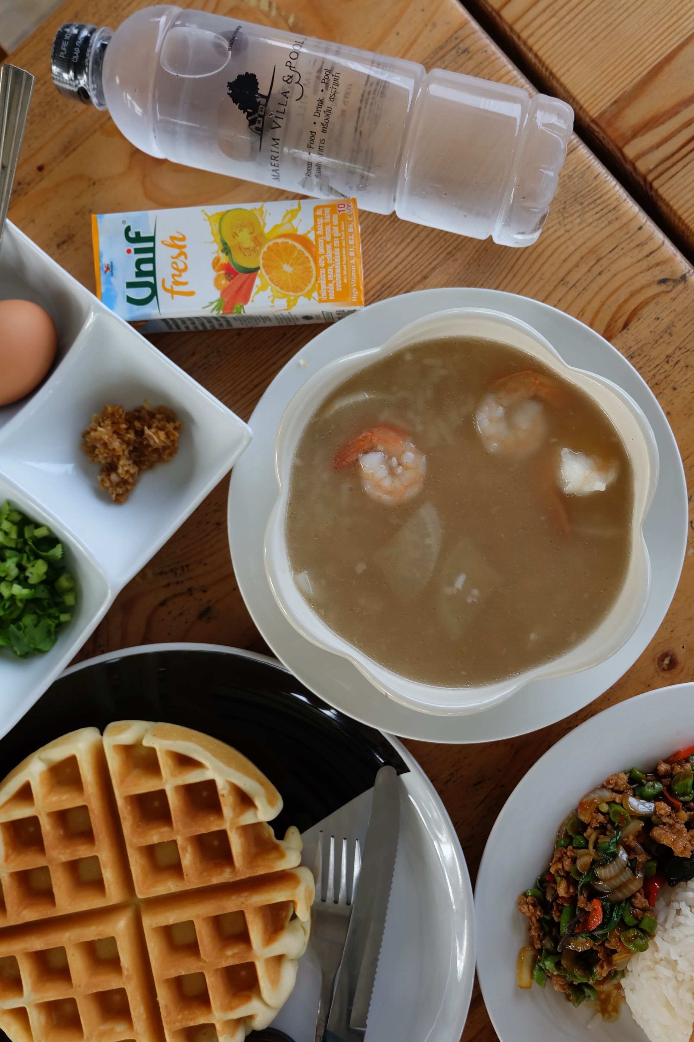 A meal with a bowl of soup, a plate of rice with vegetables, a waffle, and condiments on a wooden table.
