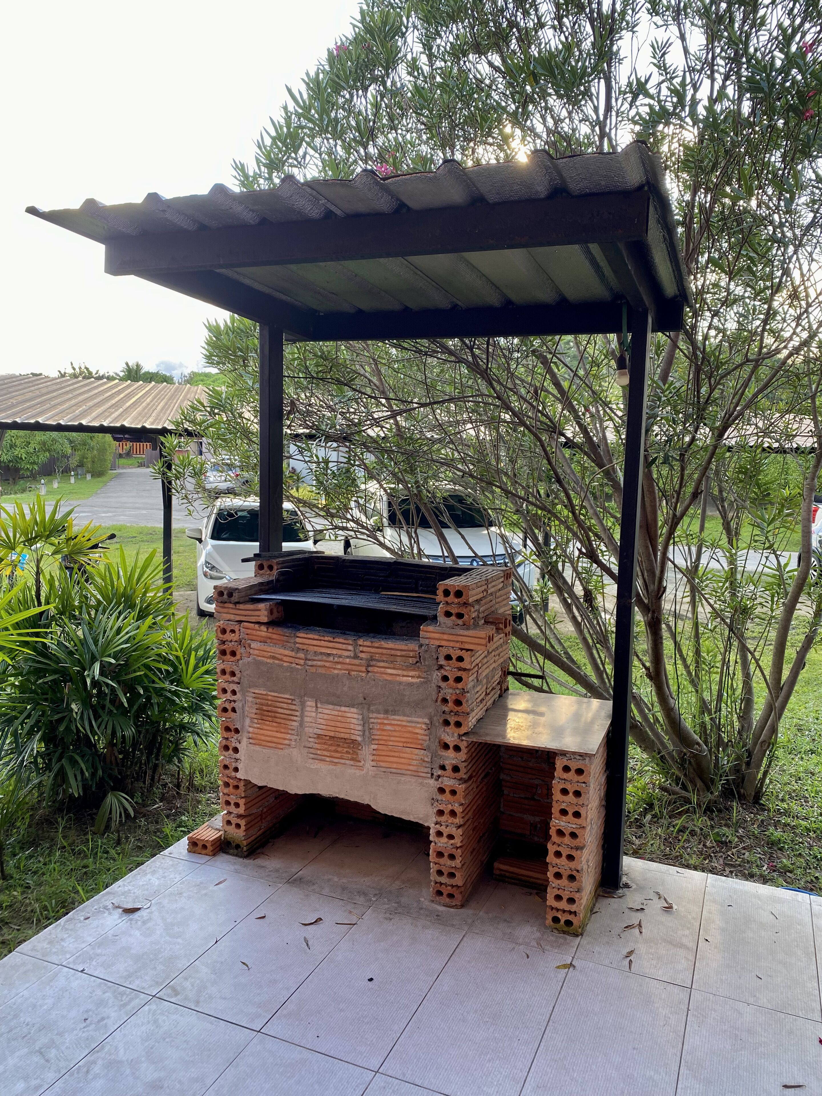 An outdoor brick barbecue grill under a metal roof, surrounded by greenery and a parked car.