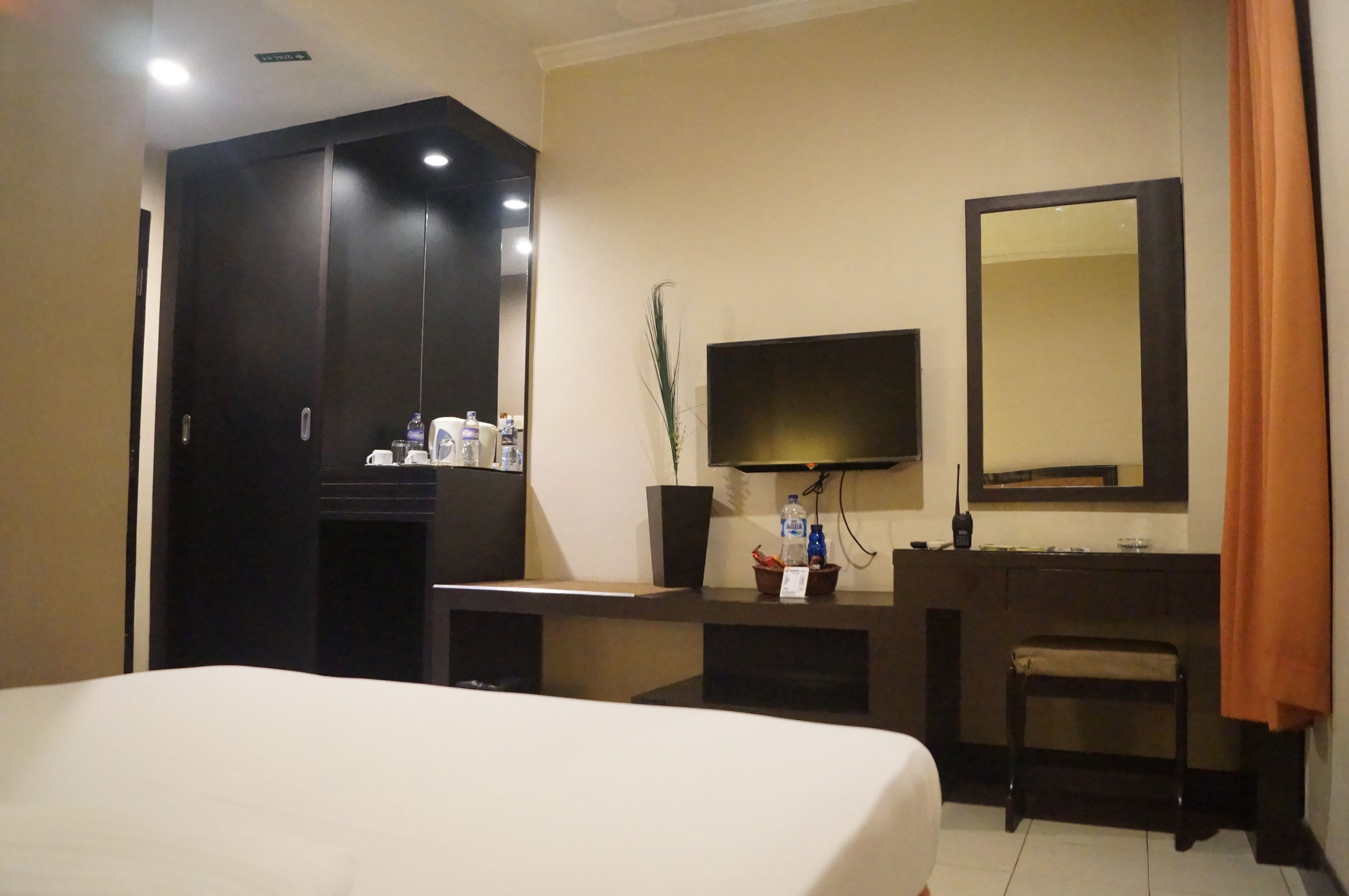 A hotel room with a bed, a dark wooden wardrobe, a desk with a flat-screen TV, a mirror, and a small stool.