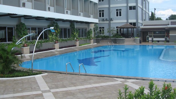 Une piscine dotée d’une terrasse carrelée, entourée de plantes en pot et bordée d’un immeuble à plusieurs étages avec des balcons.