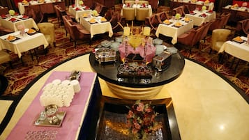 A formal dining area with multiple tables set for service, a central decorative display, and a floral arrangement on a reflective surface.