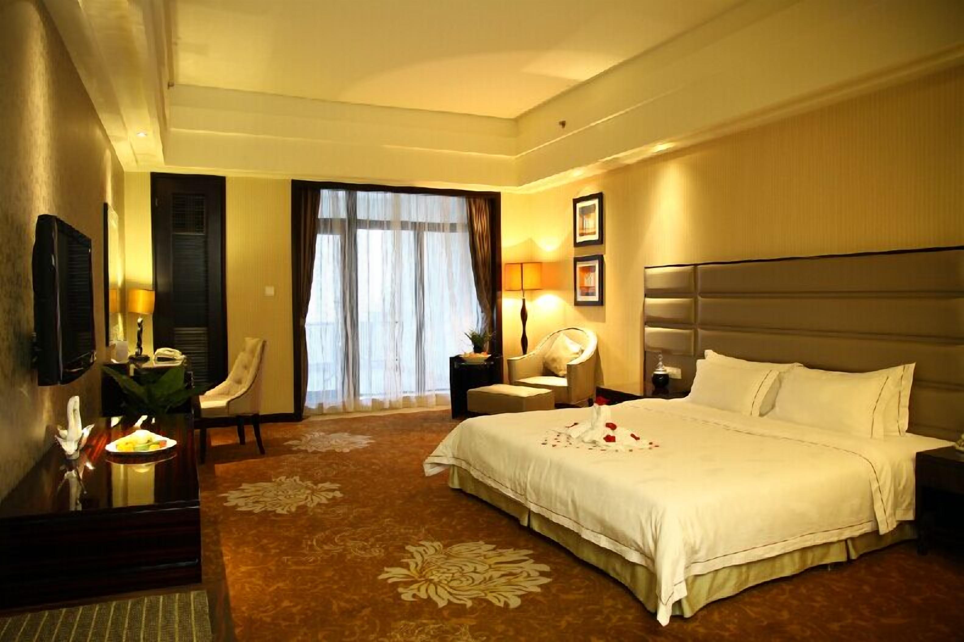 A hotel room with a large bed, a desk with a lamp, a chair, a nightstand, and a window with curtains.