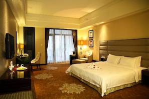 Ein Hotelzimmer mit einem großen Bett, einem Schreibtisch mit Lampe, einem Sessel, einem Nachttisch und einem Fenster mit Vorhängen.