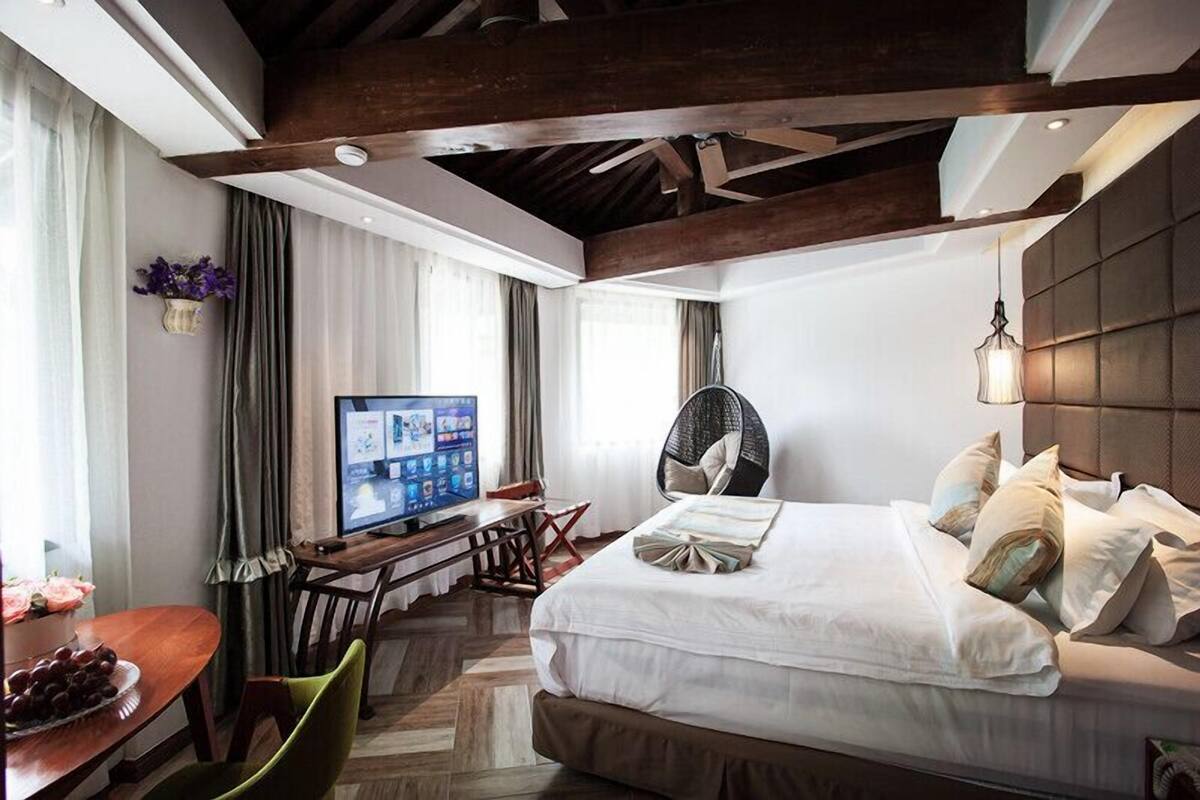 A modern hotel room with a large bed, a desk with a computer, a chair, and a dining table with fruit.