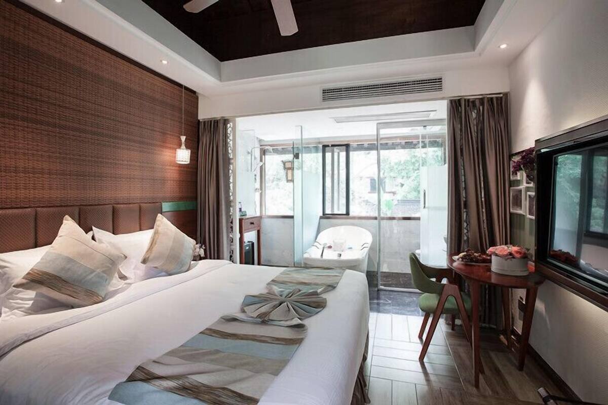 A modern hotel room with a large bed, a TV, a balcony with a chair, and a dining area with a table and chairs.