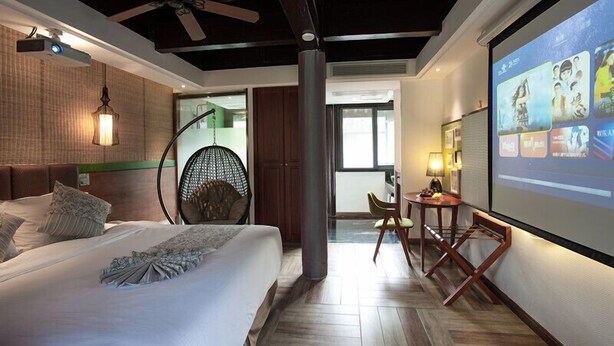 Una habitación de hotel moderna con una cama grande, ventilador de techo, pantalla de proyector mostrando imágenes y vistas al exterior.