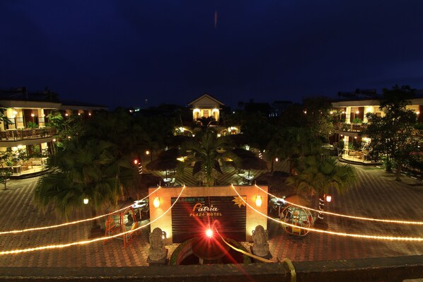 Entrada de un hotel con un letrero que dice "Patia Plaza Hotel", rodeada de palmeras e iluminada con luces colgantes.