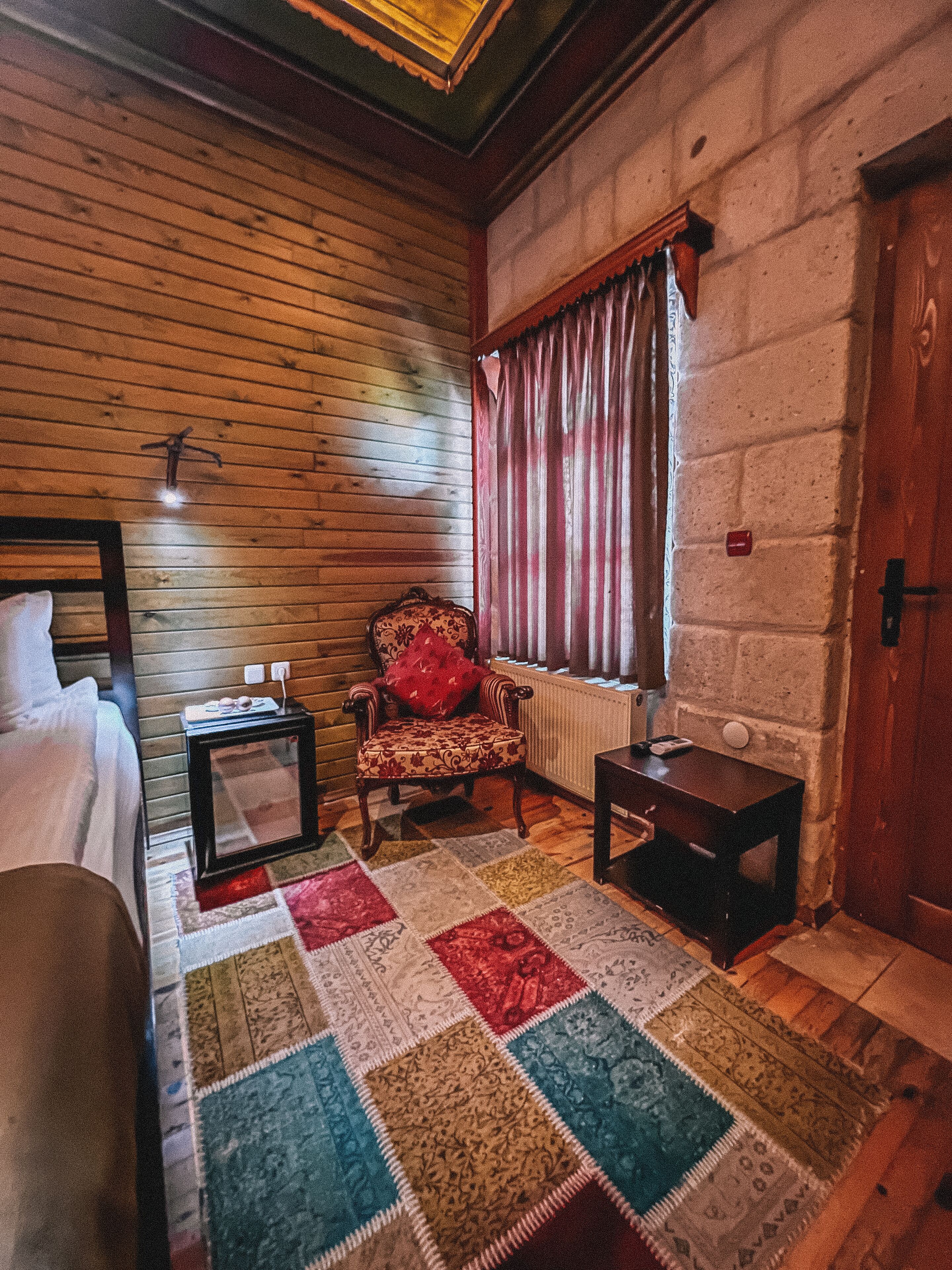 Una habitación acogedora con techo de madera, una cama, una silla con cojín rojo, una mesita y una ventana con cortinas.