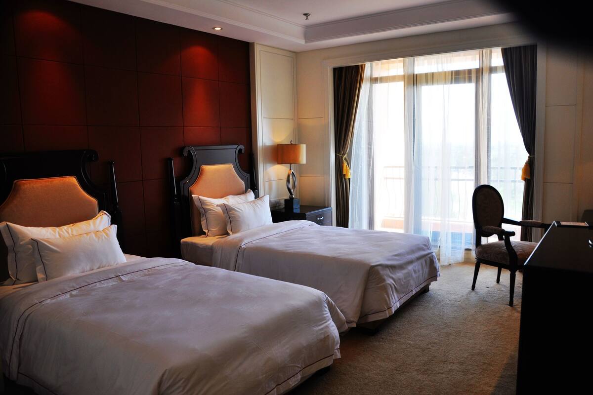 Une chambre d’hôtel avec deux lits, un bureau, une chaise, une lampe et une vue sur la ville grâce à de grandes fenêtres.
