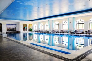 An indoor swimming pool with a clear blue ceiling, large arched windows, and sun loungers along the pool's edge.