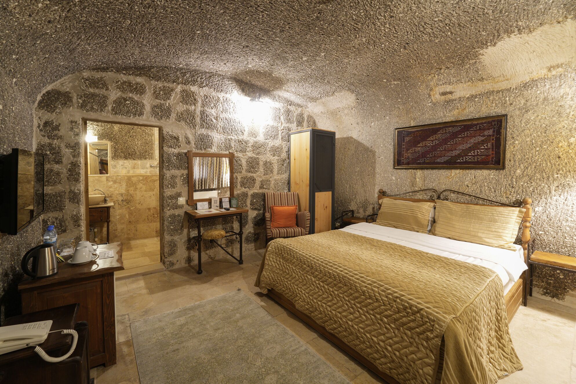 A stone-walled bedroom with a bed, a desk, a chair, a mirror, and a framed tapestry.