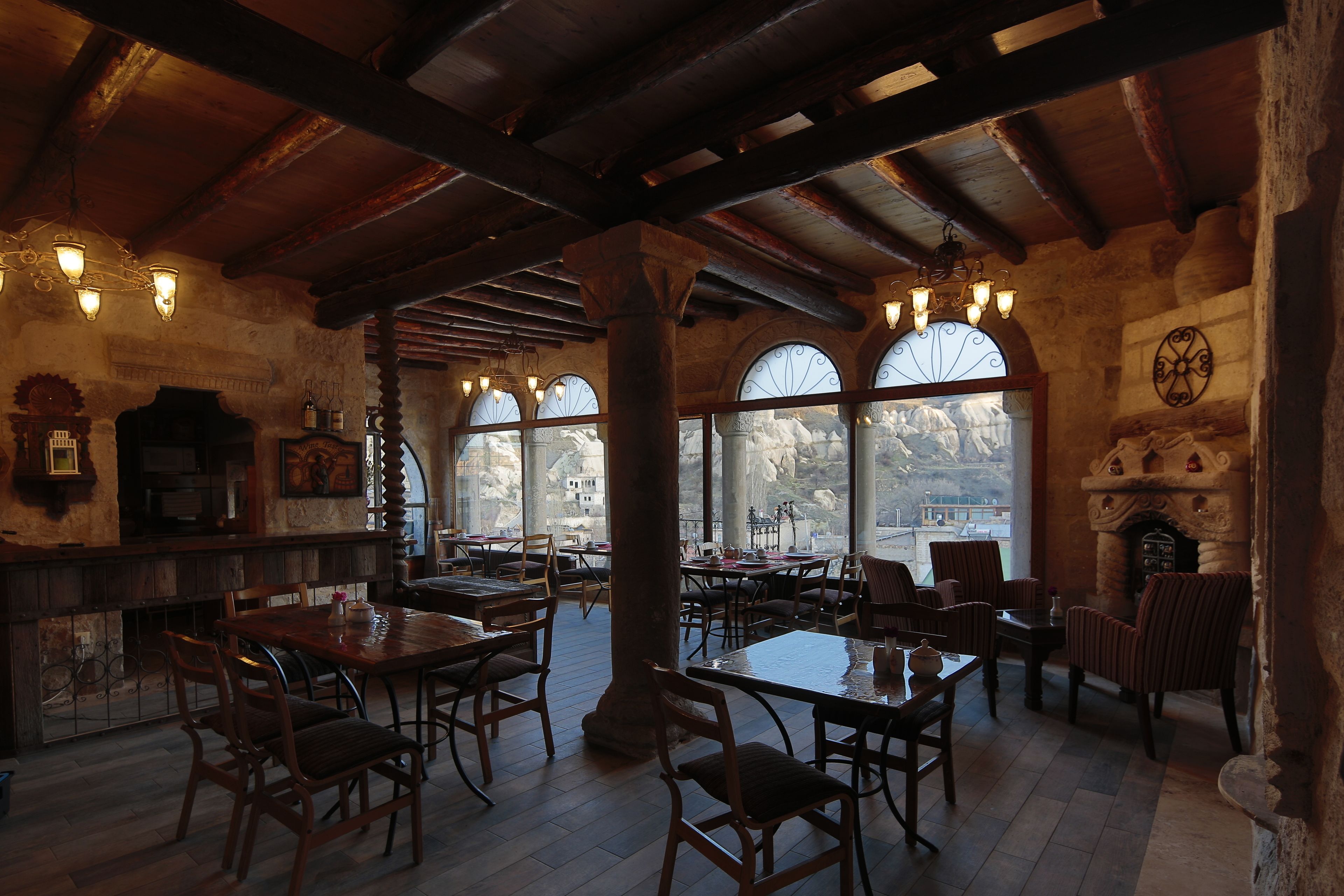 A rustic interior with wooden beams, stone walls, and arched windows. Tables and chairs are arranged for dining.