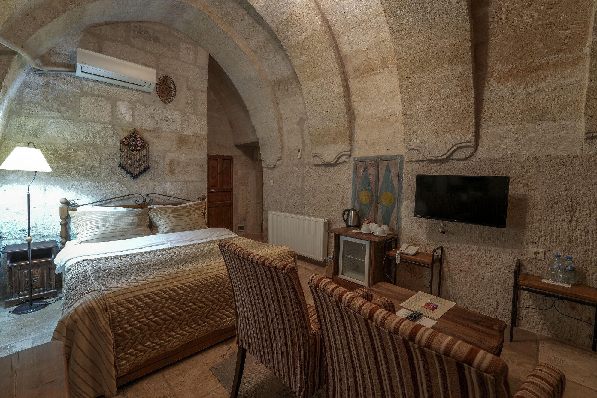 A room with a stone wall, a bed, a television, and a small table with cups and a kettle.
