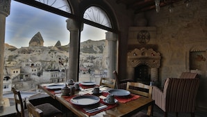 Mesa de comedor servida con platos, vasos y cubiertos, con vistas a un pueblo montañoso a través de grandes ventanas arqueadas.