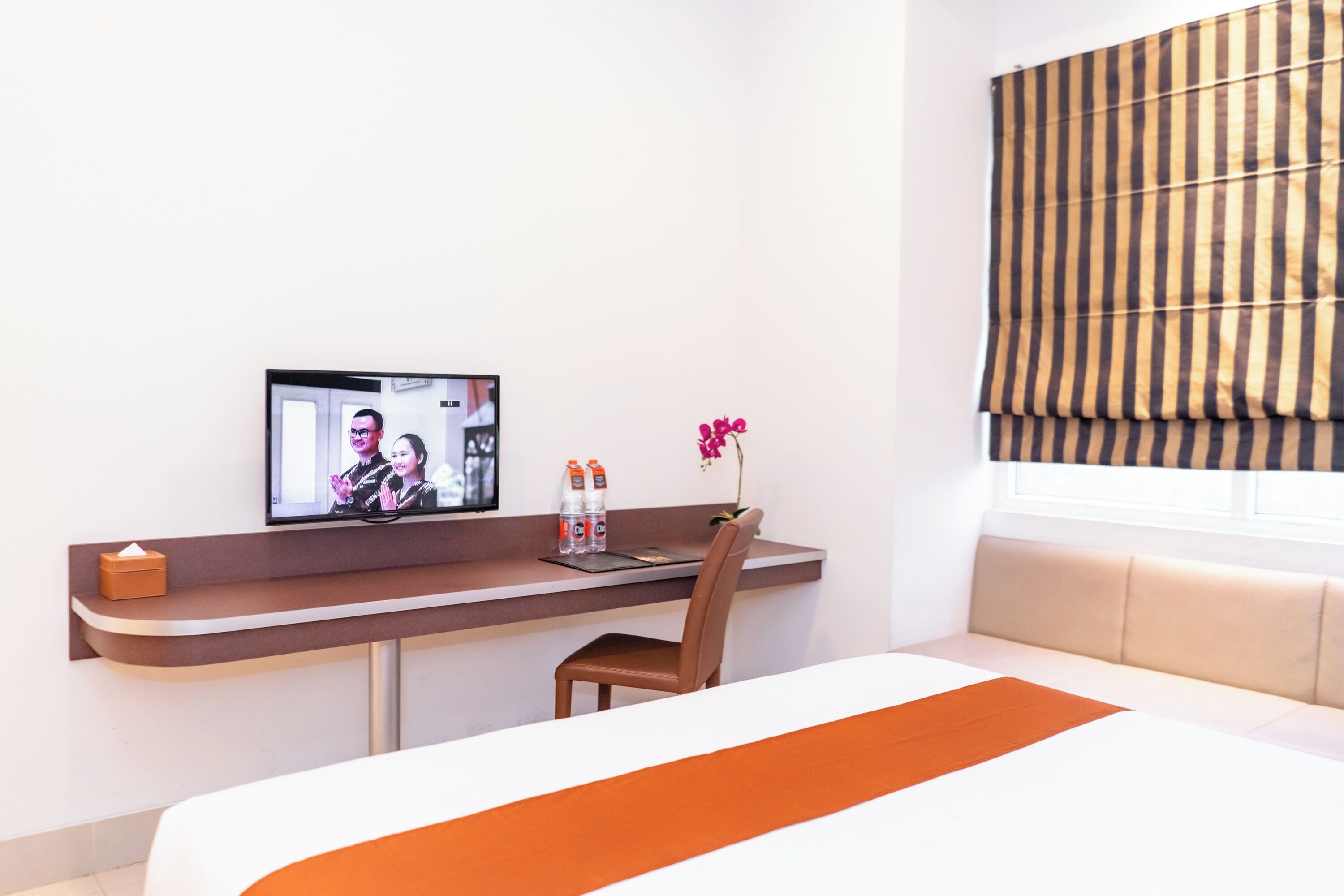 Hotelkamer met een bed, een bureau met computer, een stoel, een raam met gestreepte gordijnen en een vaas met bloemen.