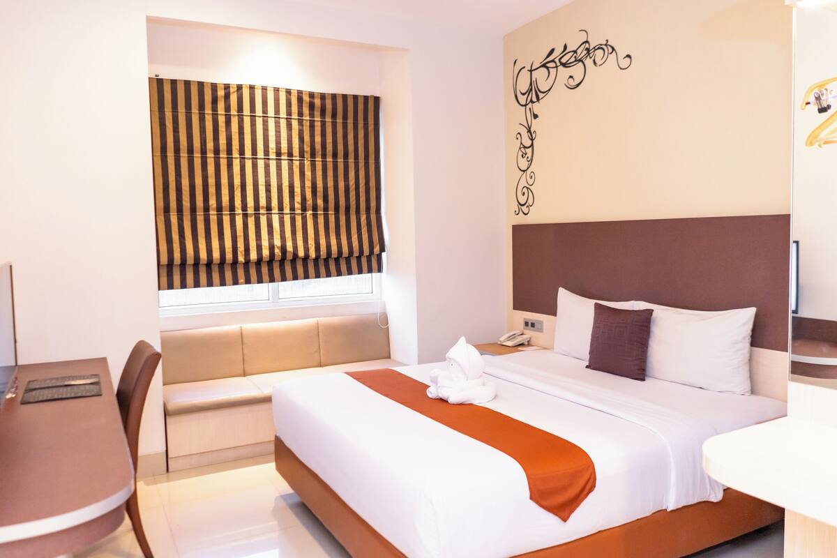 A hotel room with a bed, a desk, a chair, a window with blinds, and a wall decoration.