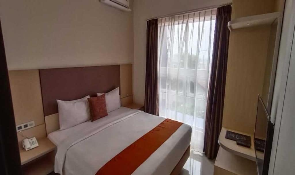 A hotel room with a bed, a bedside telephone, a window with curtains, and a wall-mounted air conditioning unit.