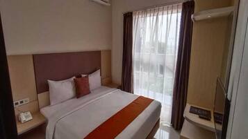 A hotel room with a bed, a bedside telephone, a window with curtains, and a wall-mounted air conditioning unit.