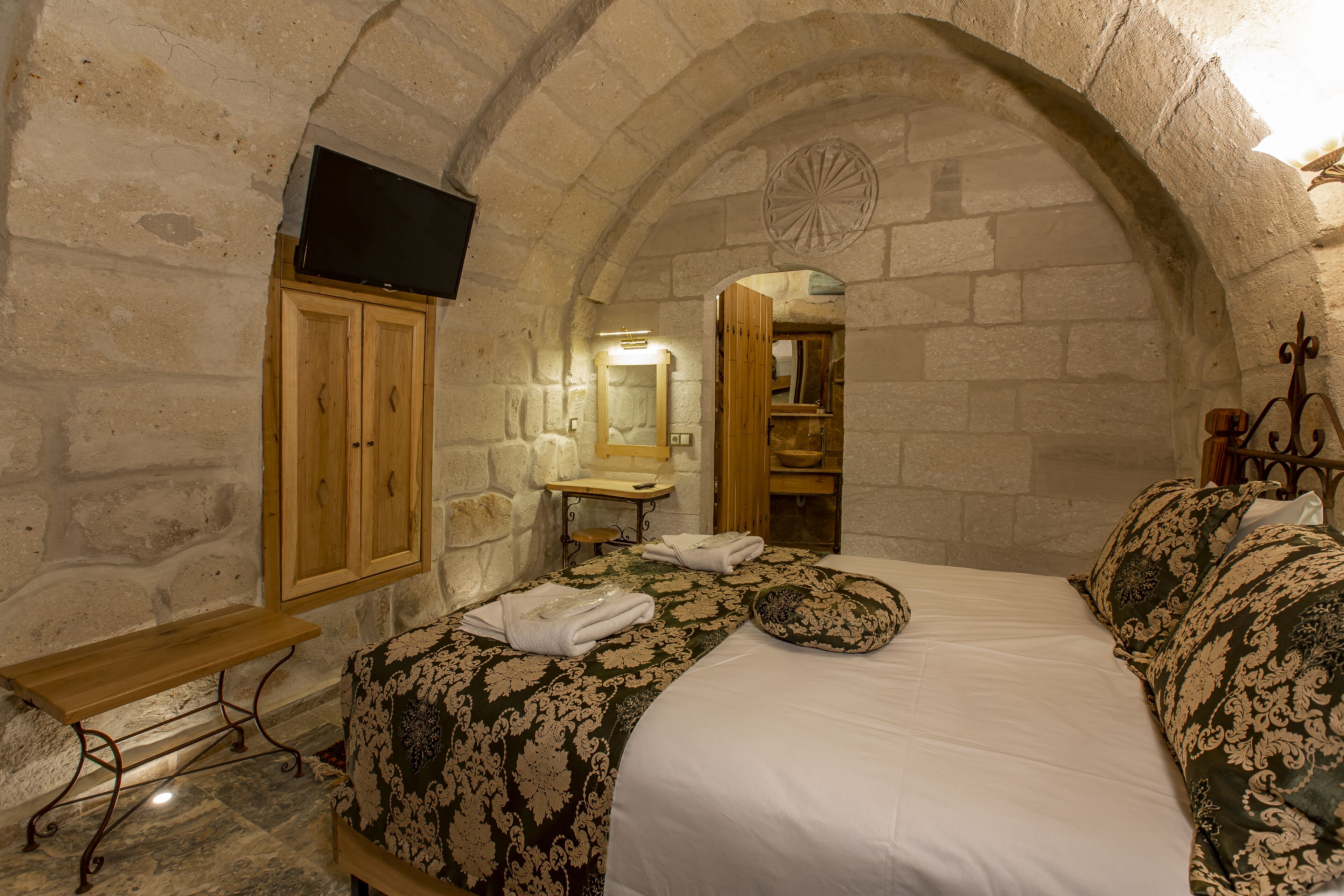 A stone-walled bedroom with a bed, wooden cabinet, and a flat-screen TV mounted on the wall.